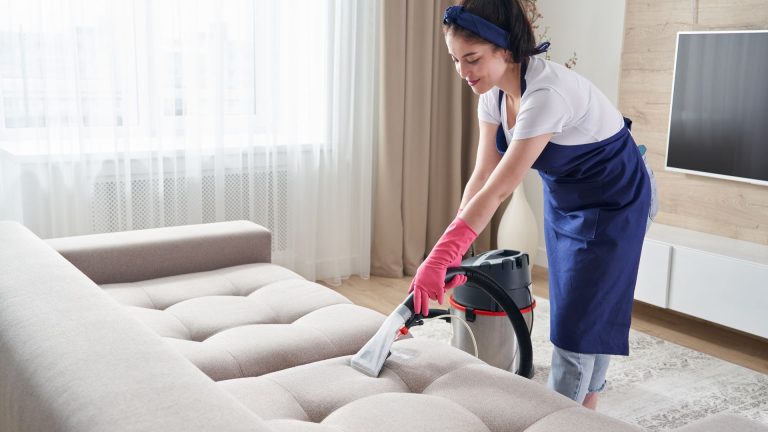 Woman cleaning couch with vacuum at home.